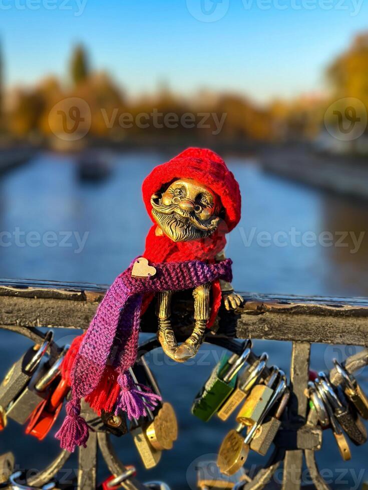 Close-up of a whimsical Homlin figurine dressed in a red hat and scarf, sitting on a bridge railing in Kaliningrad. The background features a blurred river view with vibrant autumn foliage. Captured photo