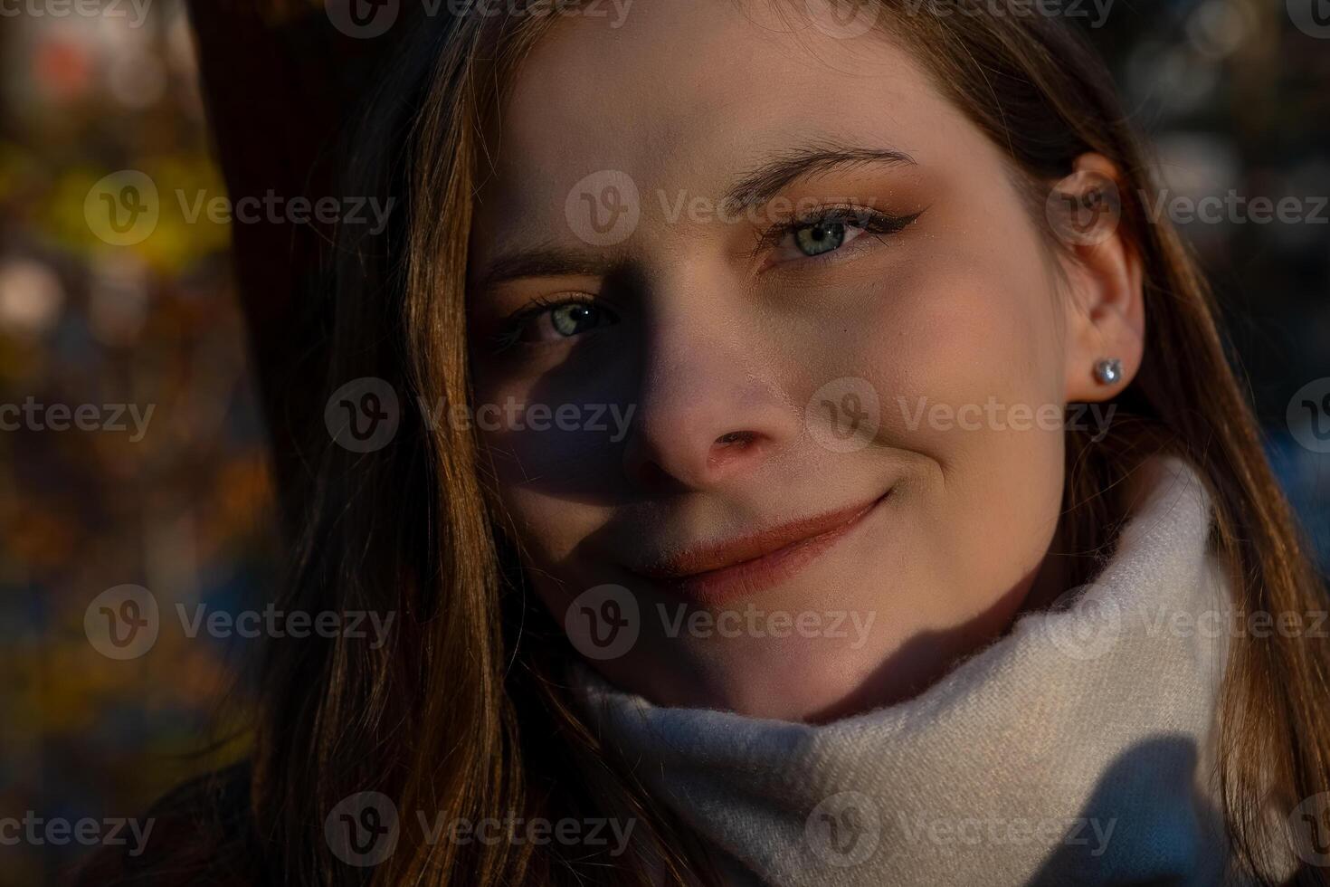 A woman with long hair smiling warmly, wearing a scarf, surrounded by autumn trees in a sunlit park during the golden hour photo