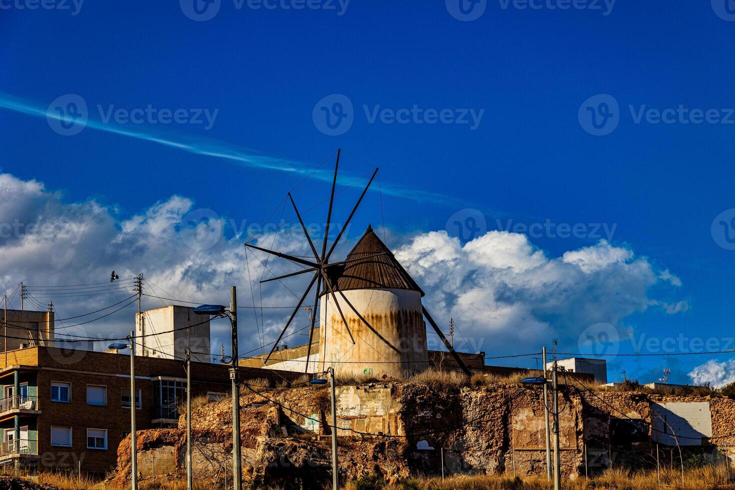 A rustic windmill stands amid old brick buildings under a bright blue sky with fluffy clouds photo