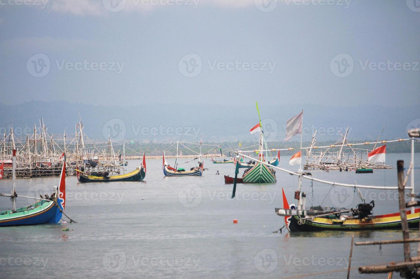 A tranquil scene of traditional fishing boats with Indonesian flags, set against a calm sea and hazy mountain horizon in the distance photo