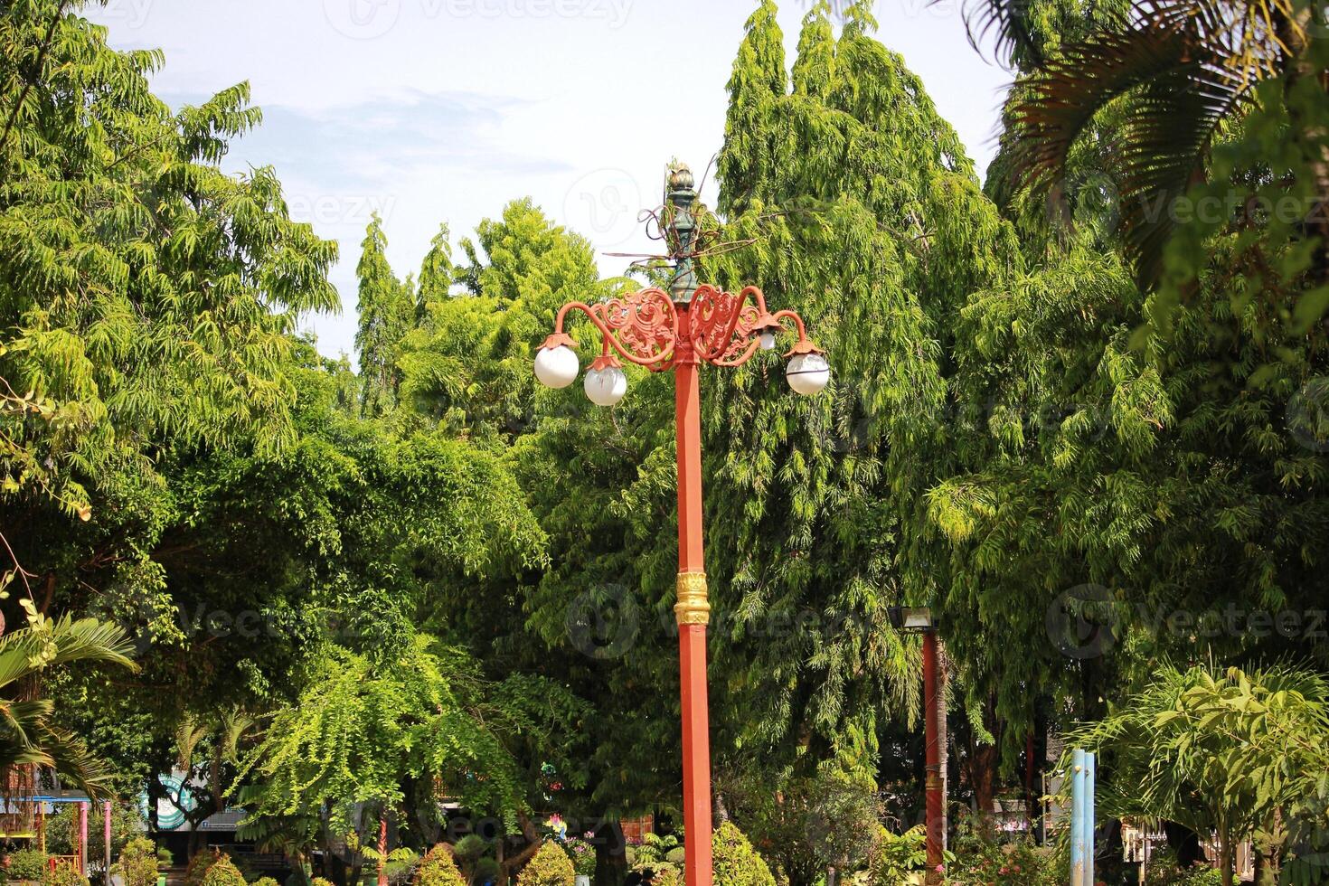A vintage red lamp post stands amidst dense green trees in an urban park photo
