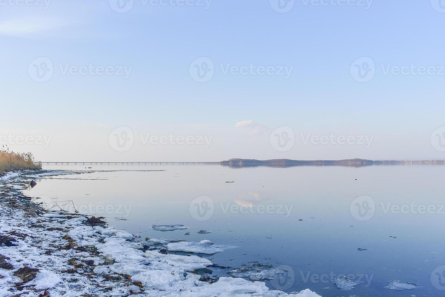 Sea shore covered with melting snow and a mirror-like surface of water reflecting the sky and trees photo