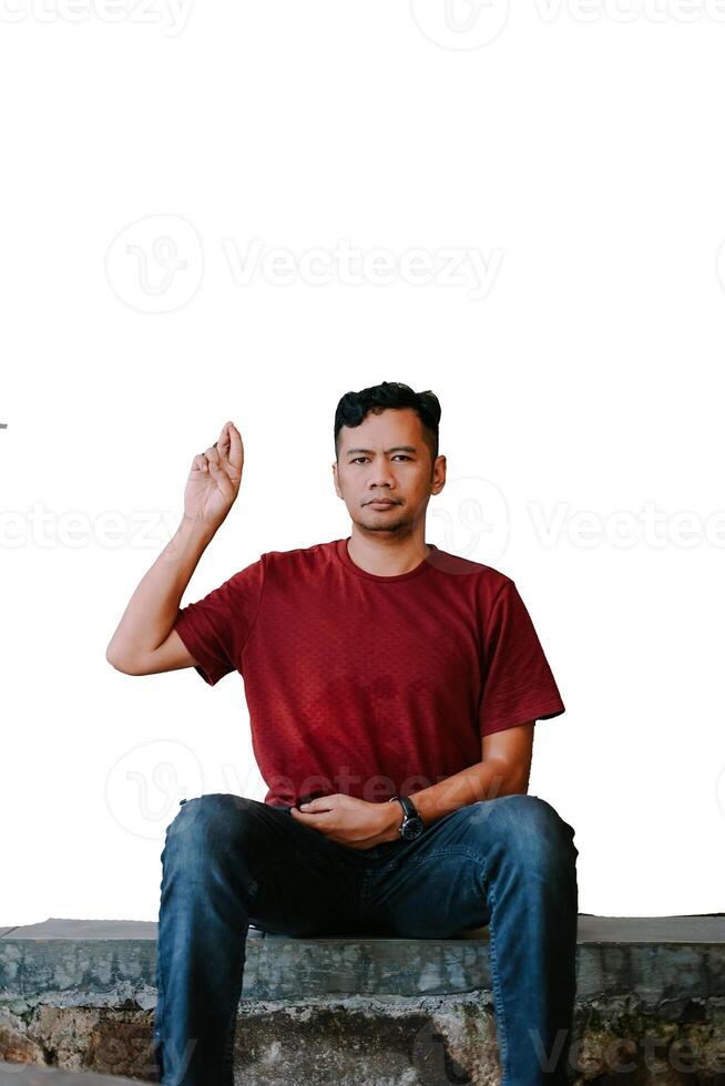A man in a red T-shirt sits on a stone ledge, raising his hand with a serious expression photo