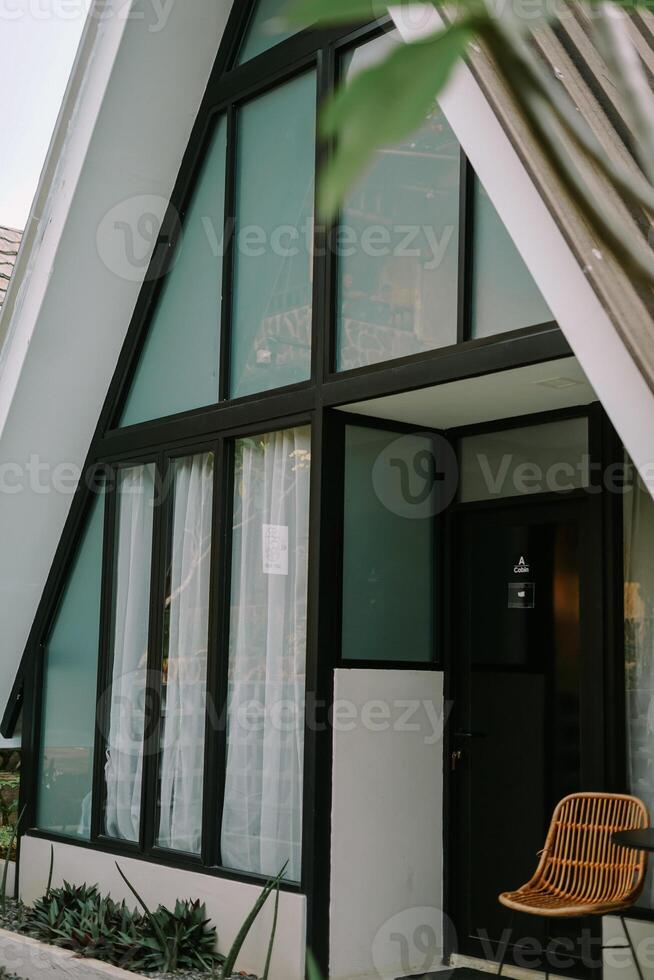 Modern A-frame cabin with large windows and a wicker chair in a cozy outdoor setting. photo