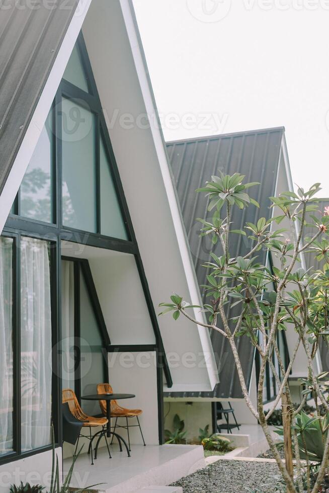 Modern A-frame cabin with large windows and a wicker chair in a cozy outdoor setting. photo
