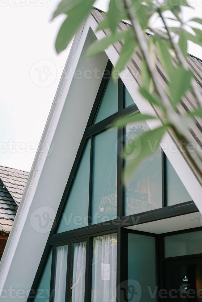 Modern A-frame cabin with large windows and a wicker chair in a cozy outdoor setting. photo