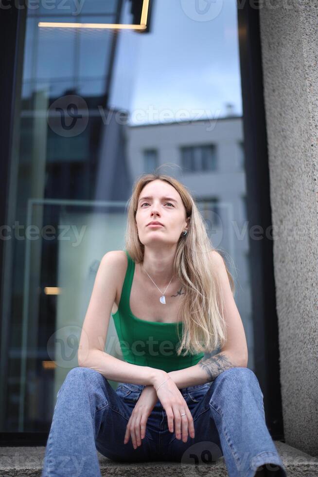 Young Woman in Green Tank Top Sitting Outside a Modern Building in Daylight photo