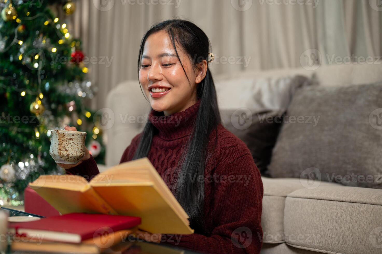 A happy woman sits in her living room, enjoying a book and some snacks during the Christmas holiday. photo
