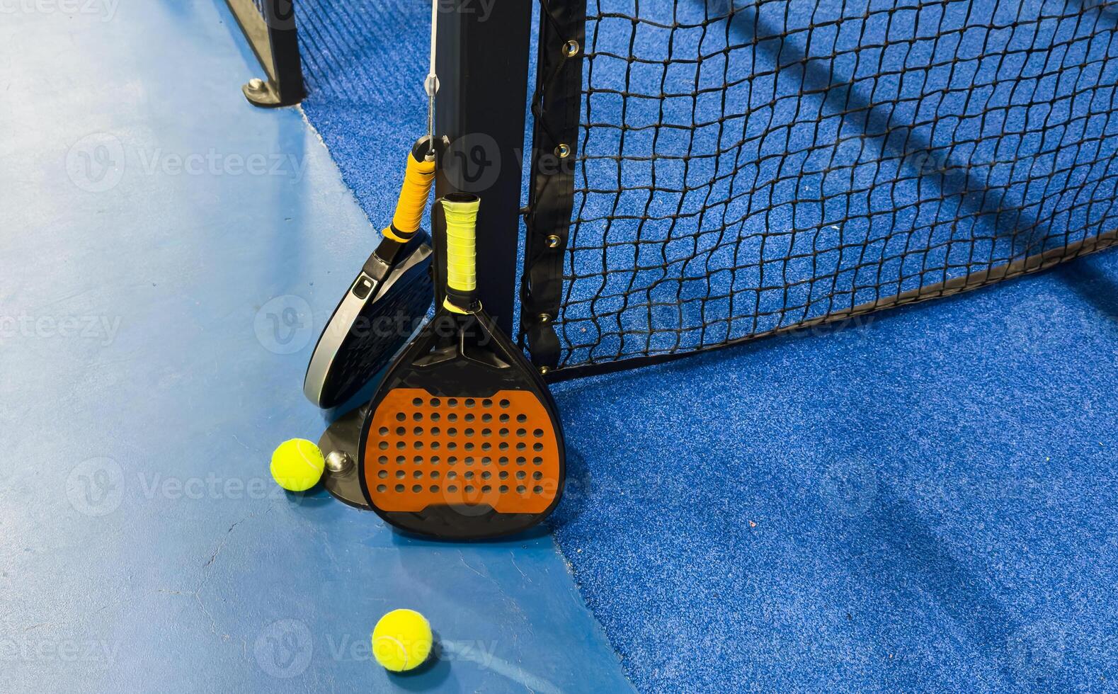 Photo of an orange padel racket and a ball on the ground of a blue outdoor court. Padel concept. Paddle equipment.