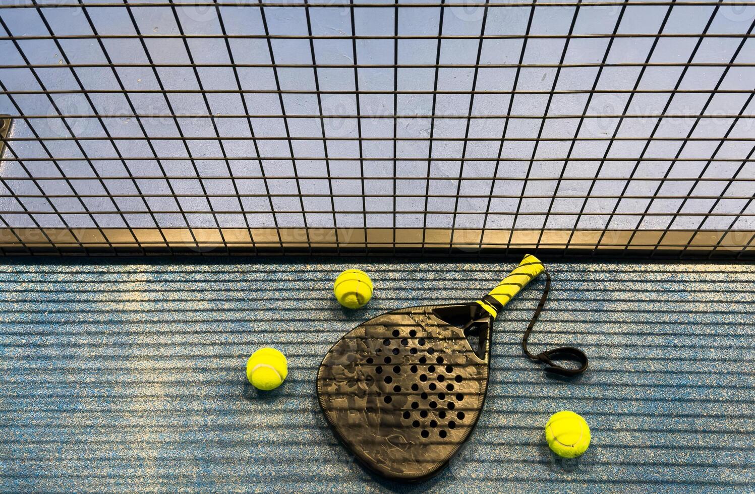 A padel or paddle tennis racket and a bright yellow tennis ball lie on a blue court. The net and a blurred figure of a player are visible in the background. photo