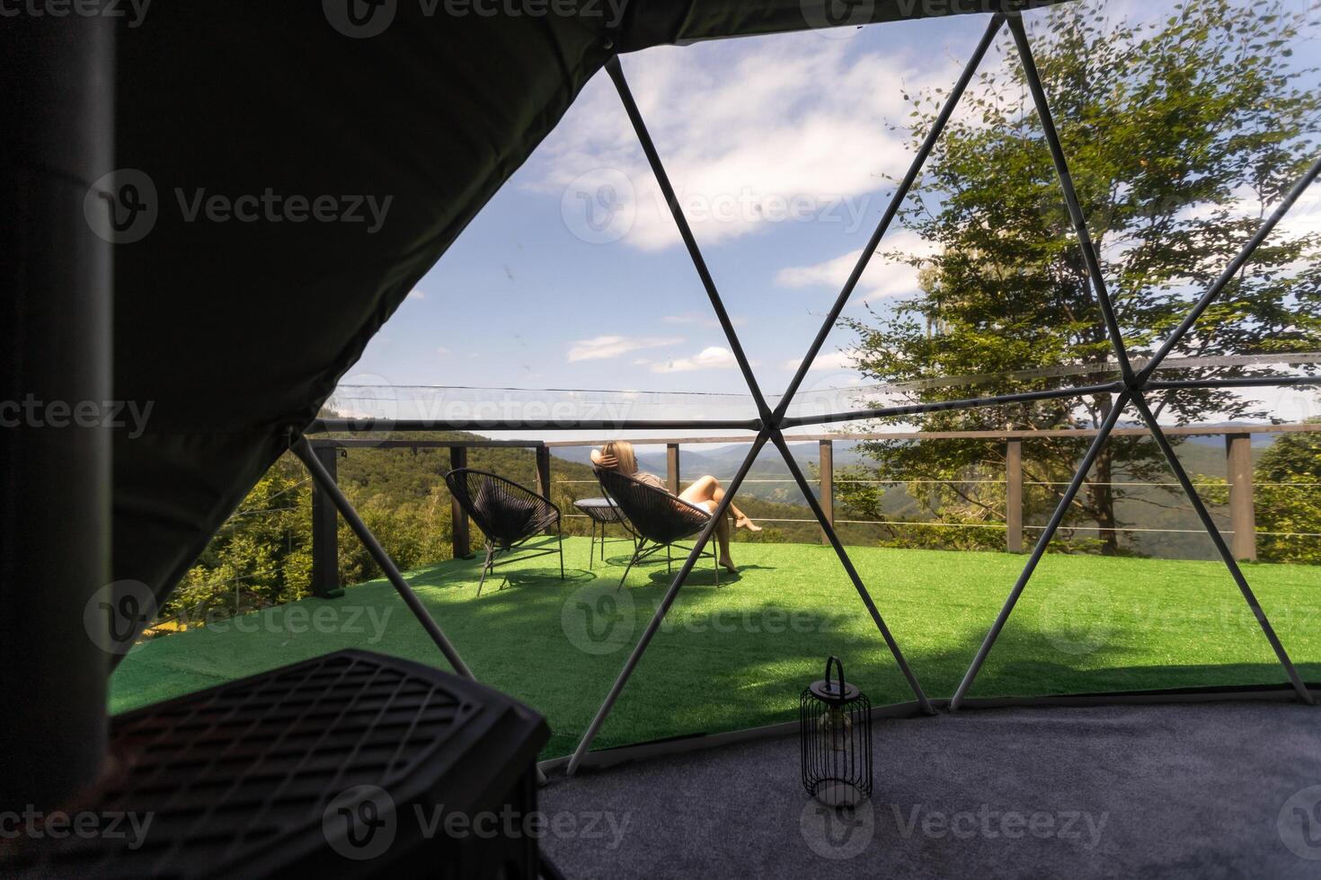 Young woman resting on sunbed on the wooden terrace near the modern house with panoramic windows near pine forest. Concept of solitude and recreation on nature photo