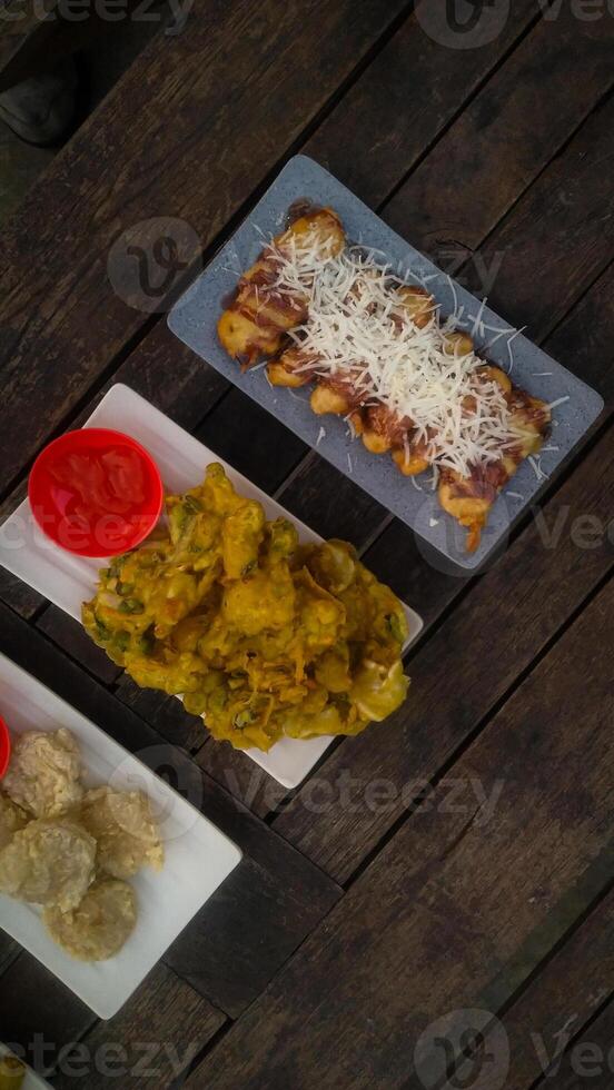 A top-down view of an assortment of Indonesian street food served on a rustic wooden table. A fried banana with grated cheese on top, a plate of vegetable fritters. photo
