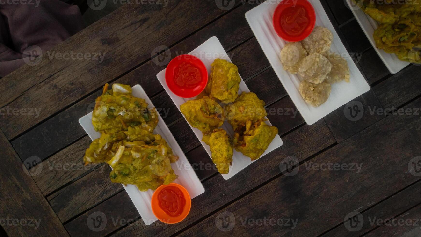 A top-down view of an assortment of Indonesian street food served on a rustic wooden table. A fried banana with grated cheese on top, a plate of vegetable fritters. photo