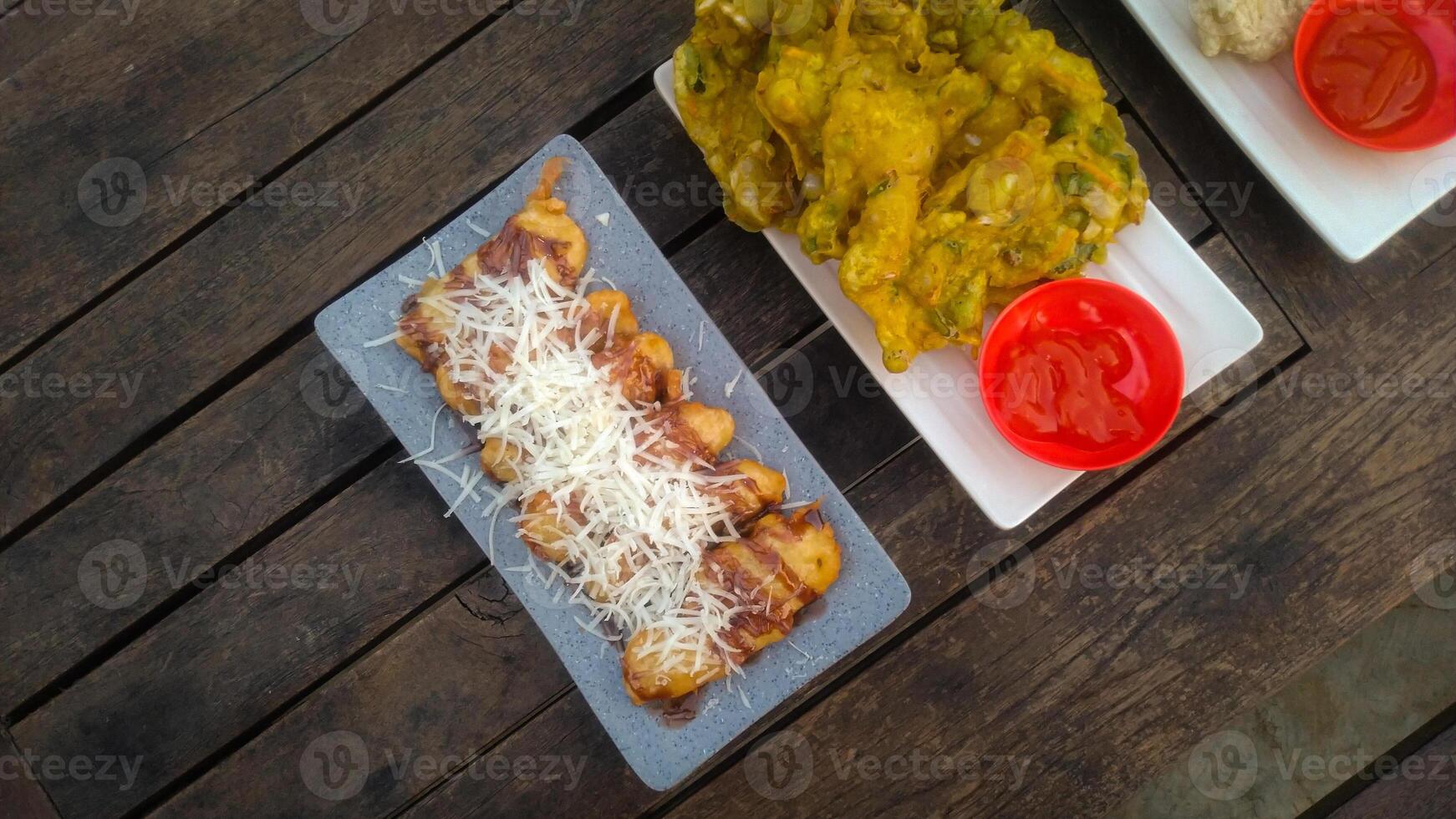 A top-down view of an assortment of Indonesian street food served on a rustic wooden table. A fried banana with grated cheese on top, a plate of vegetable fritters. photo