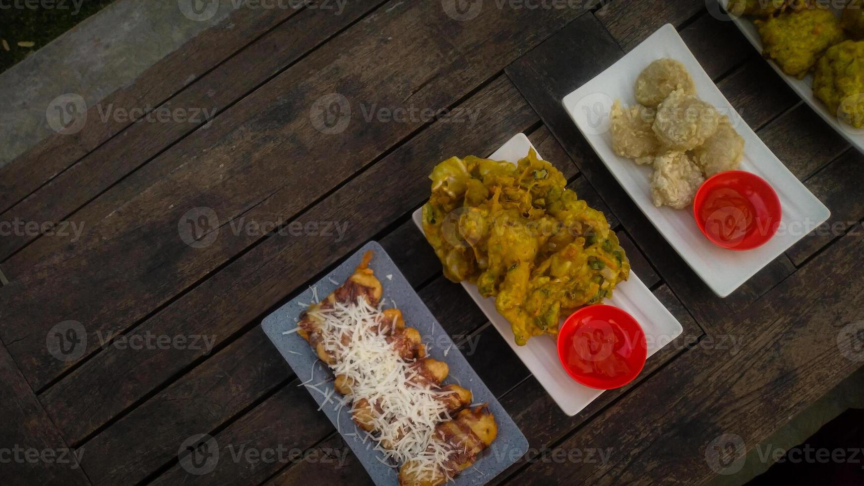 A top-down view of an assortment of Indonesian street food served on a rustic wooden table. A fried banana with grated cheese on top, a plate of vegetable fritters. photo