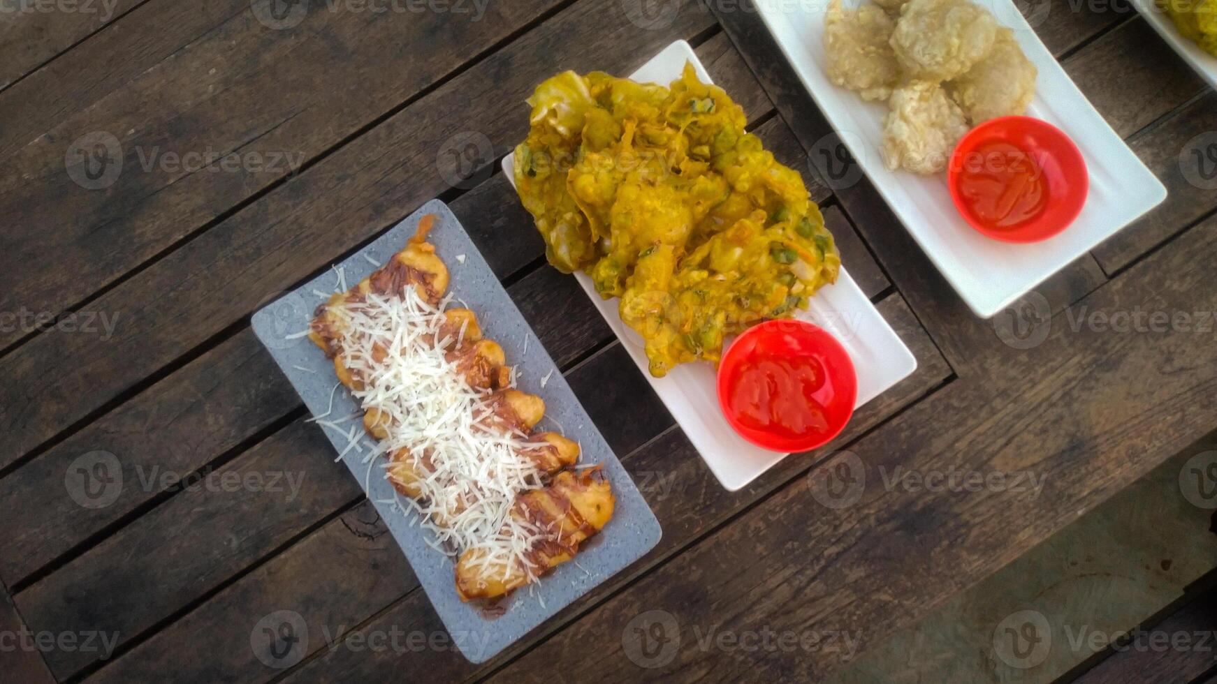 A top-down view of an assortment of Indonesian street food served on a rustic wooden table. A fried banana with grated cheese on top, a plate of vegetable fritters. photo