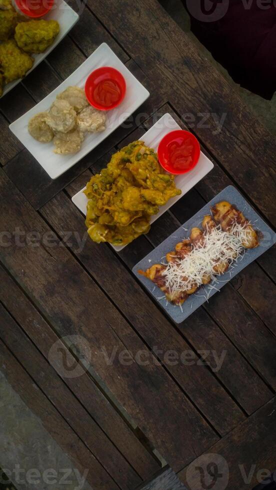 A top-down view of an assortment of Indonesian street food served on a rustic wooden table. A fried banana with grated cheese on top, a plate of vegetable fritters. photo