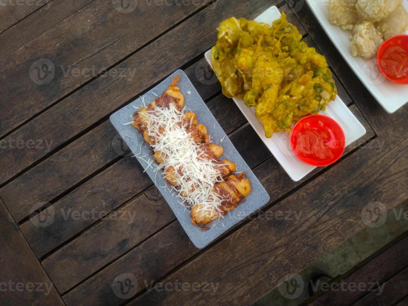 A top-down view of an assortment of Indonesian street food served on a rustic wooden table. A fried banana with grated cheese on top, a plate of vegetable fritters. photo