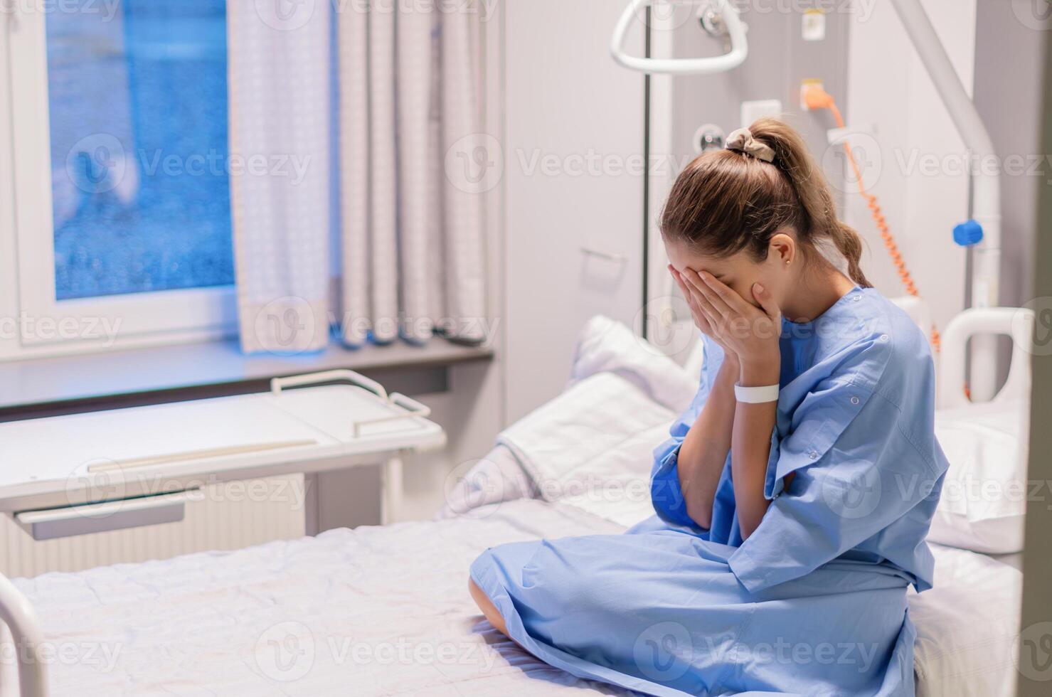 young woman in a blue hospital gown sits alone on a bed in a private room in a clinic, expressing her concern about the results of an examination, covering her face with both palms, photo