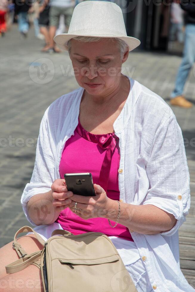 sixty year old caucasian woman in a hat and stylish summer clothes chats in a smartphone on a bench in the city, her backpack lies nearby, photo