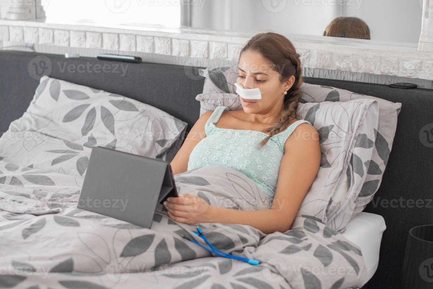 Woman resting in bed in casual clothes and bandages on her face after plastic surgery on her nose,using tablet to communicate with family, calm atmosphere and soft colors illustrate recovery process photo