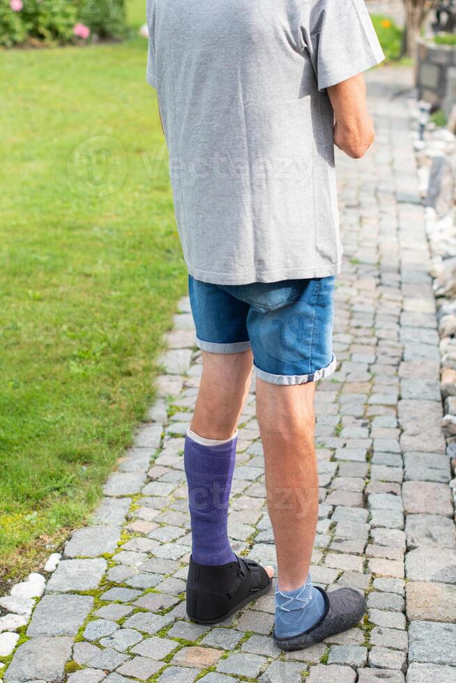 middle-aged man walks along a rocky path with a broken leg in a purple fibreglass cast, his foot shod in a special walking sandal,concept of an active lifestyle despite the limitations of an injury, photo