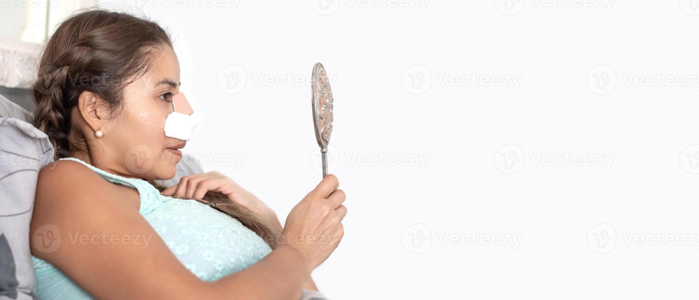 woman lies in bed in casual clothes and with bandages on her face after plastic surgery on her nose and looks at her reflection in the mirror, worried and anxious about the result,copy space photo