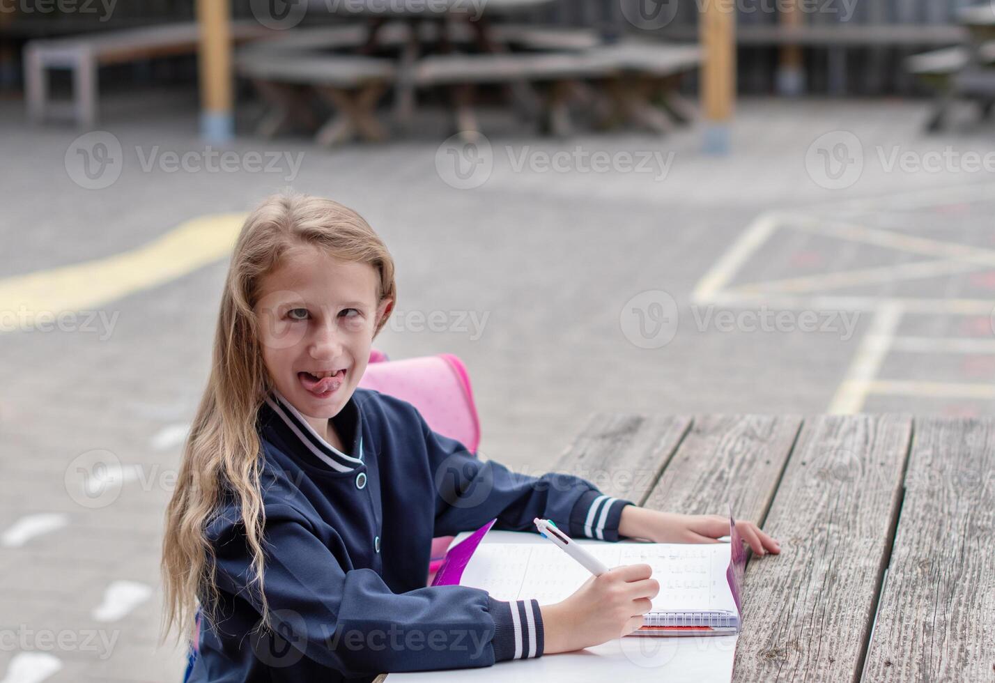 naughty girl making funny faces after successfully completing homework, at wooden table on playground, education concept, back to school, girl in beautiful blue uniform with pink backpack, photo