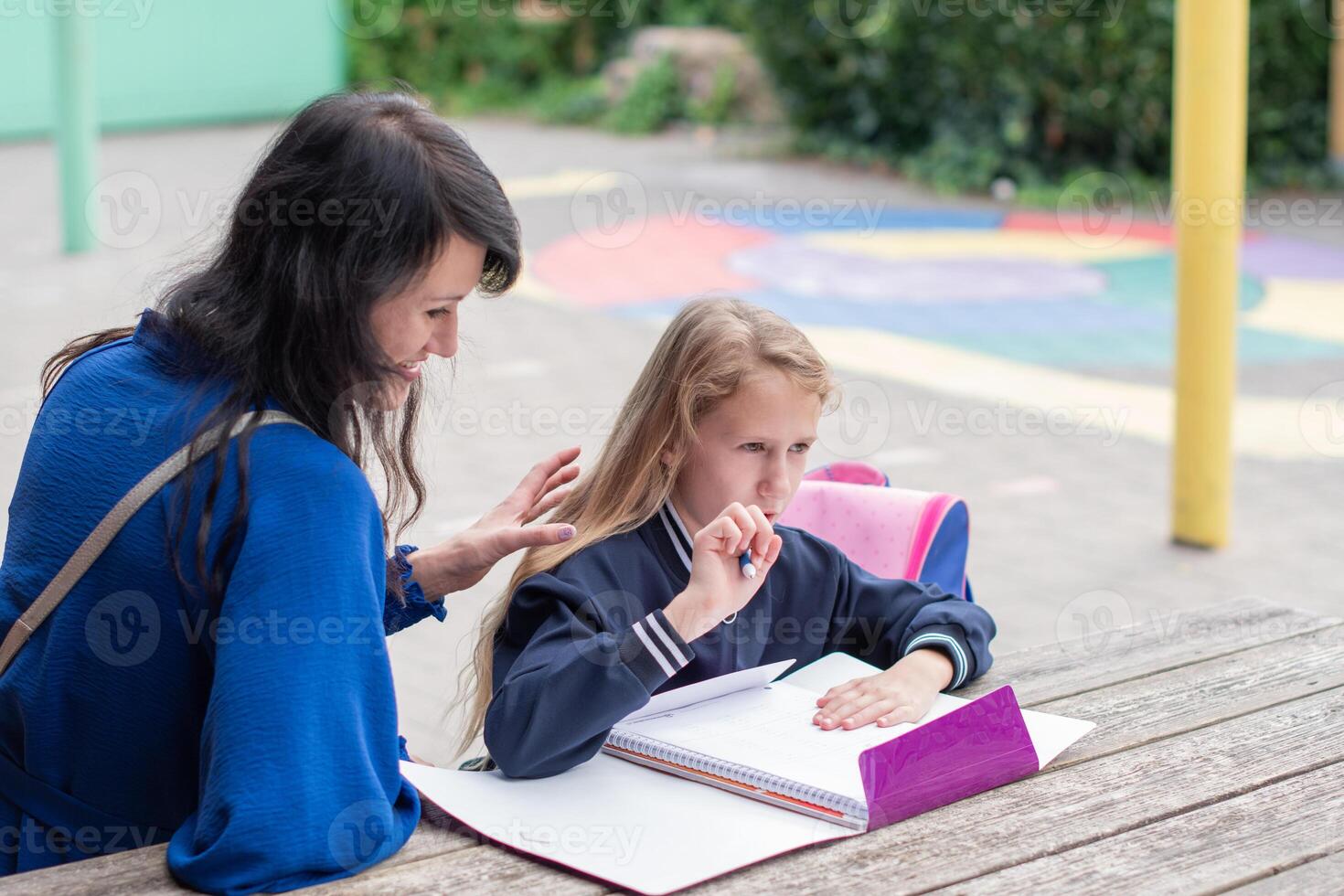 smart girl solving math problems on her own while her mother sits nearby at wooden table on children's playground and watches,education concept,going back to school, girl in beautiful blue uniform photo