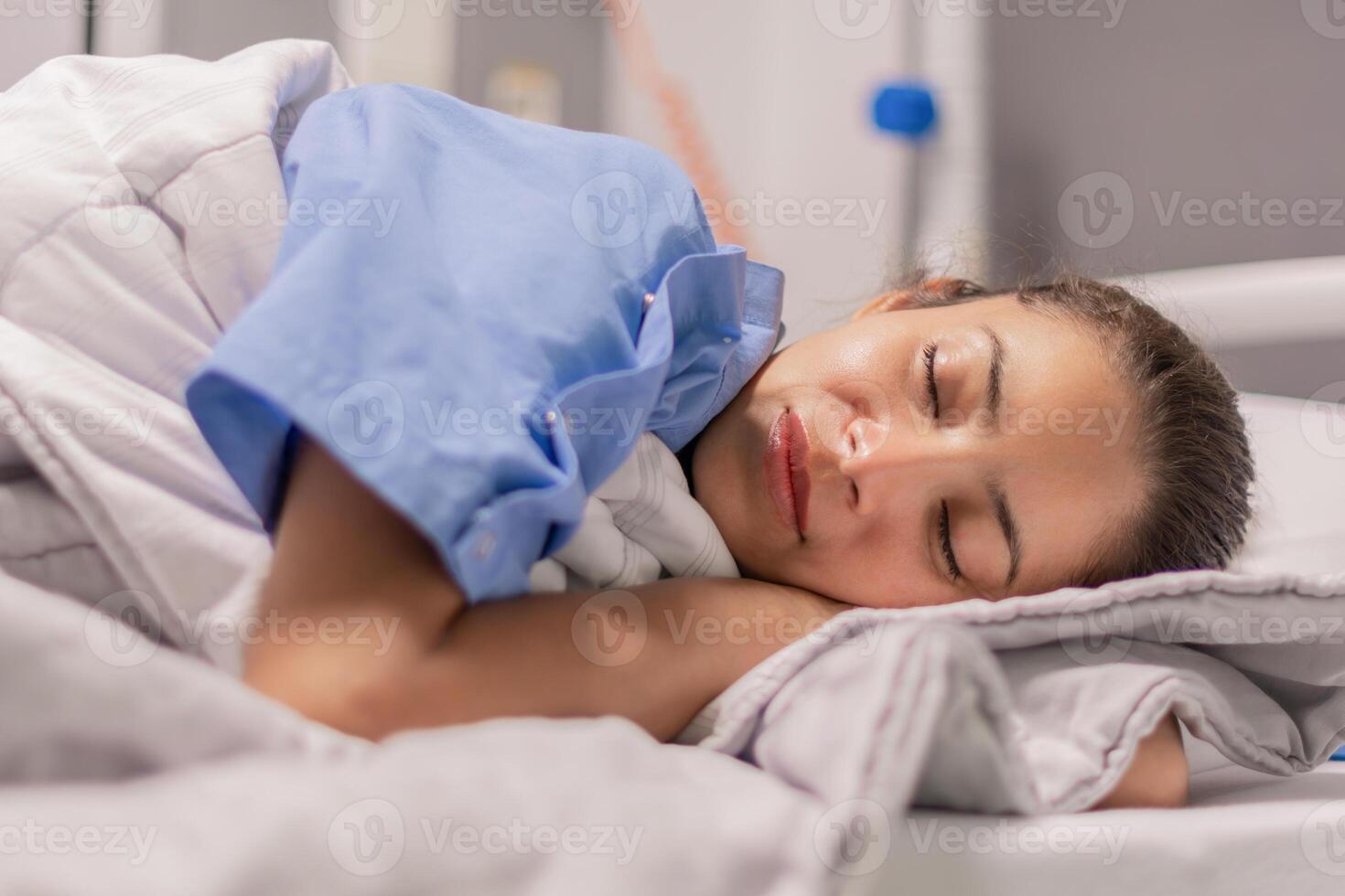 Young woman in blue robe lying comfortably and getting ready for bed on hospital bed in calm environment of clinic in private room, soft lighting, concept of recovery and care, photo