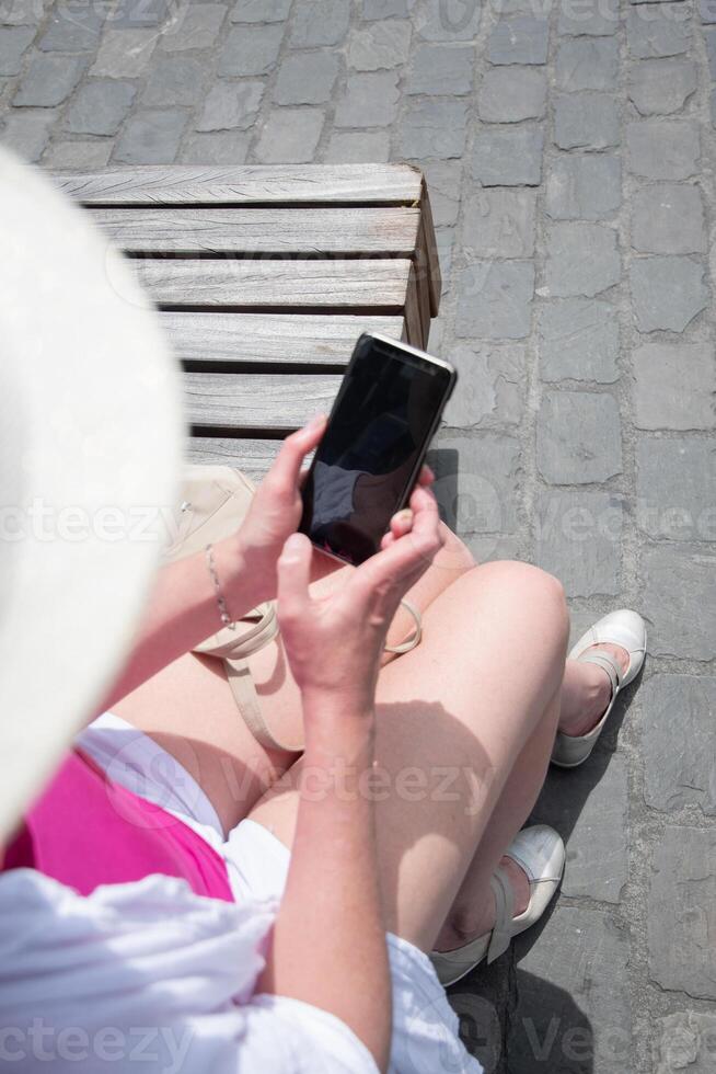 Sixty year old Caucasian woman in hat and stylish summer clothes chatting on smartphone on bench in city, rear and top view, photo