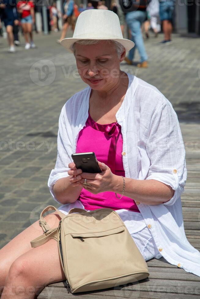 sixty year old caucasian woman in a hat and stylish summer clothes chats in a smartphone on a bench in the city, her backpack lies nearby, photo