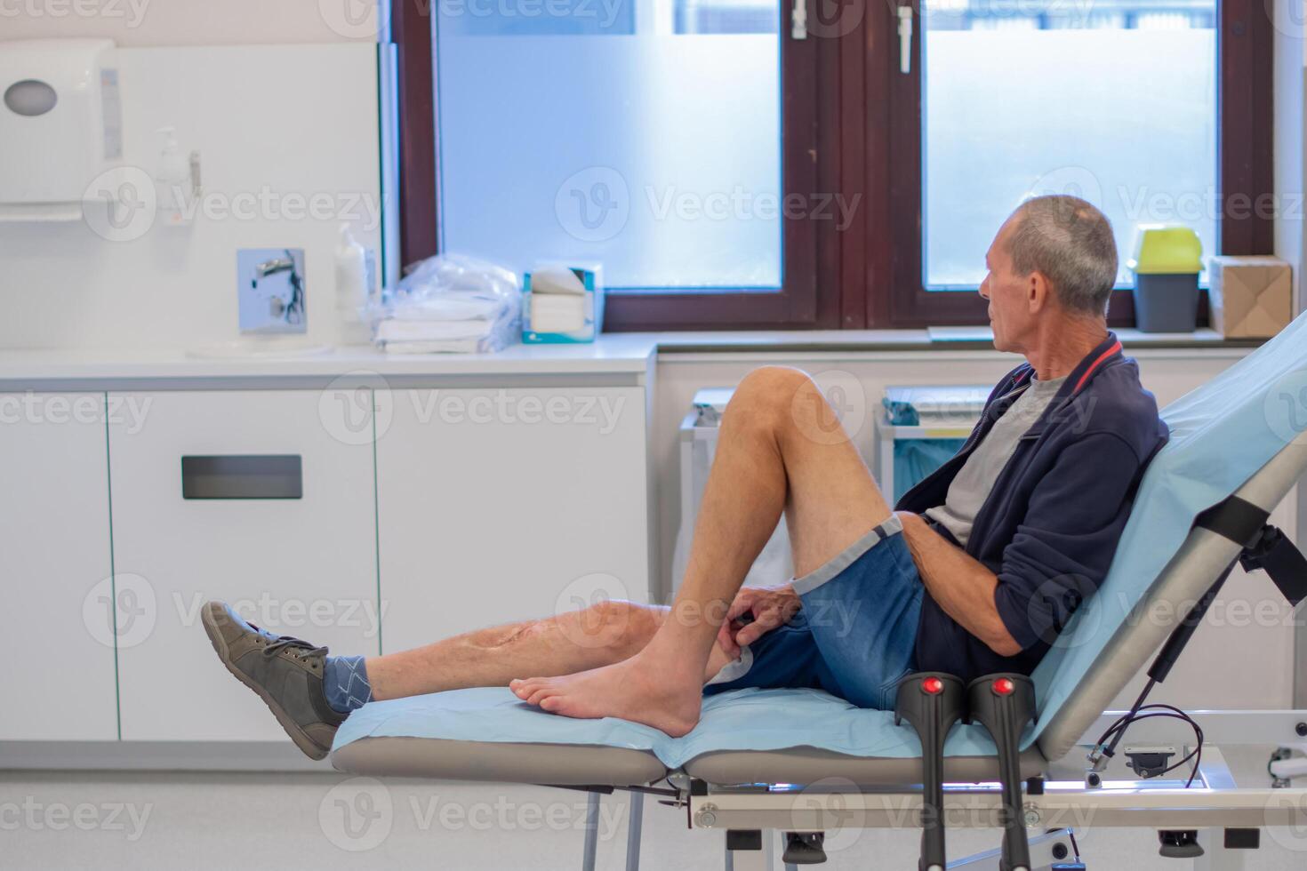 middle aged man sits deep in thought on a medical couch in a clinic office, waiting for the doctor to examine him and put a cast on his broken leg, professional first aid concept, photo
