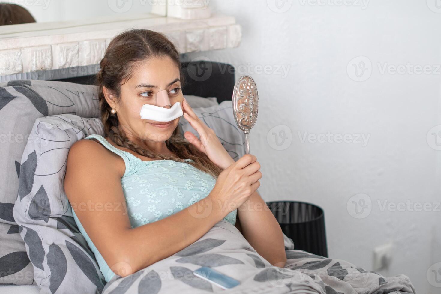 woman lies in bed in casual clothes and with bandages on her face after plastic surgery on her nose and looks at her reflection in the mirror, worried and anxious about the result, photo