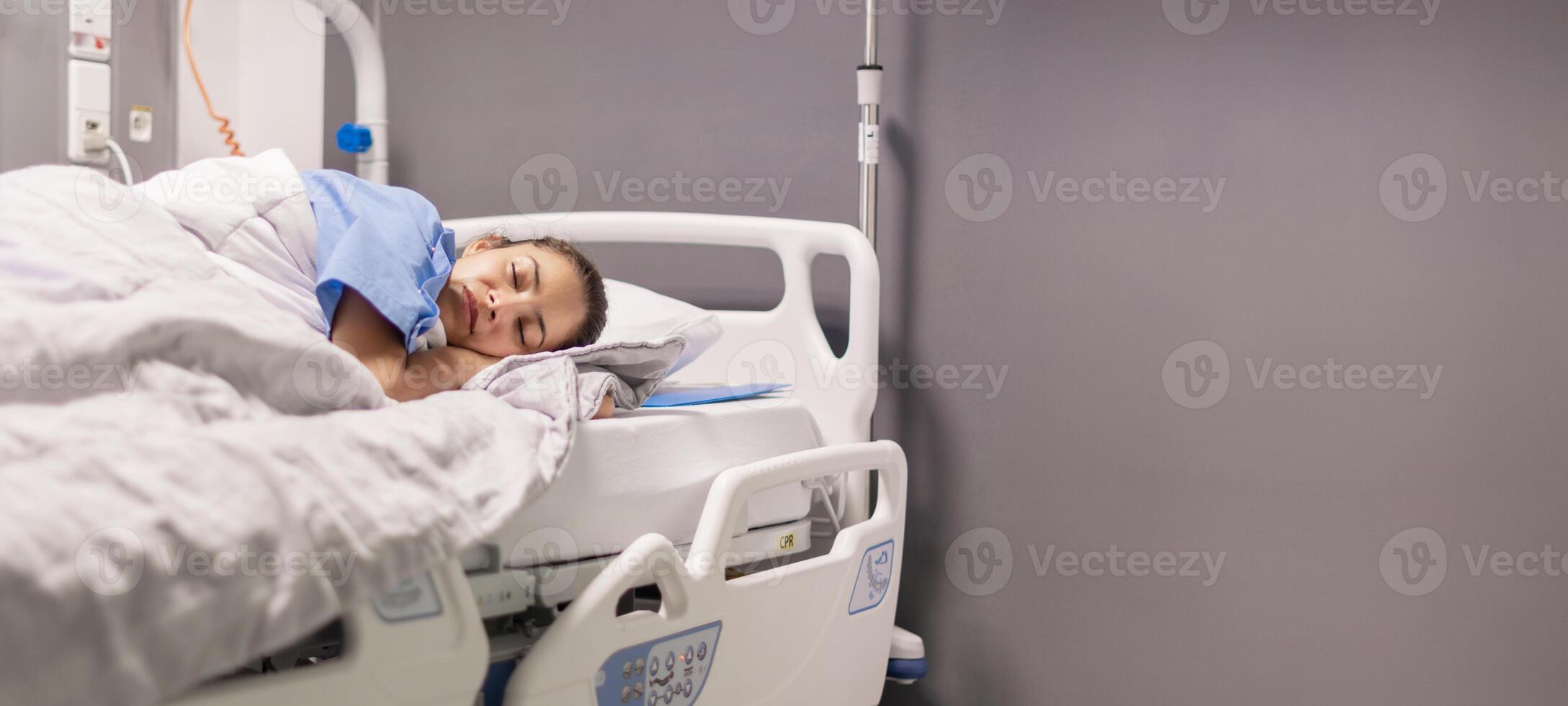 Young female patient of the clinic in blue robe sleeping on hospital bed in calm environment in private room, soft lighting, concept of recovery and care, copy space photo