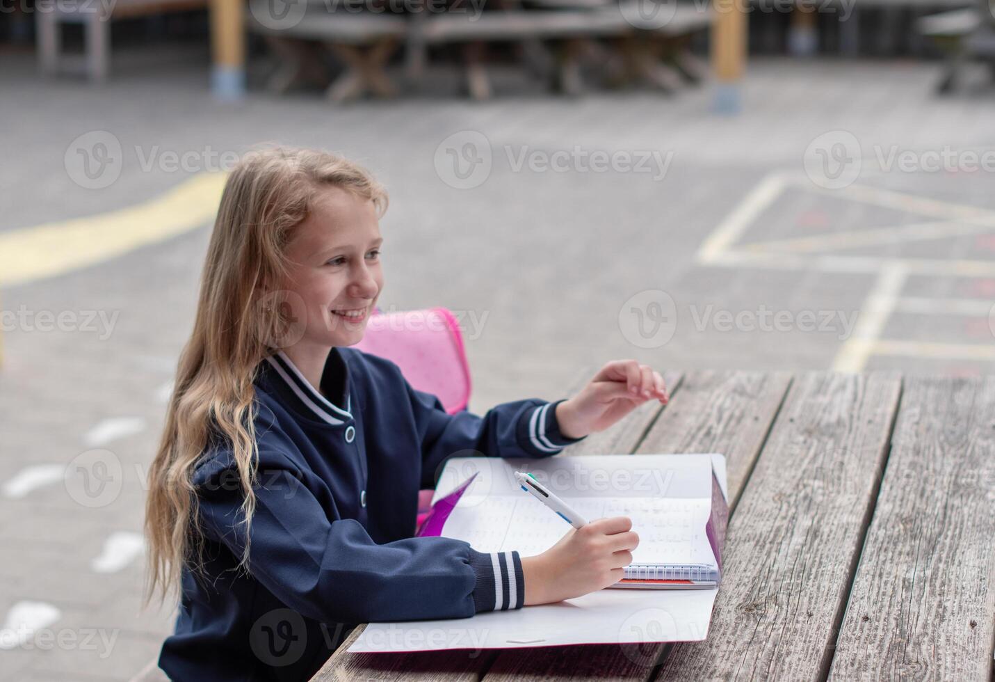 naughty girl making funny faces after successfully completing homework, at wooden table on playground, education concept, back to school, girl in beautiful blue uniform with pink backpack, photo