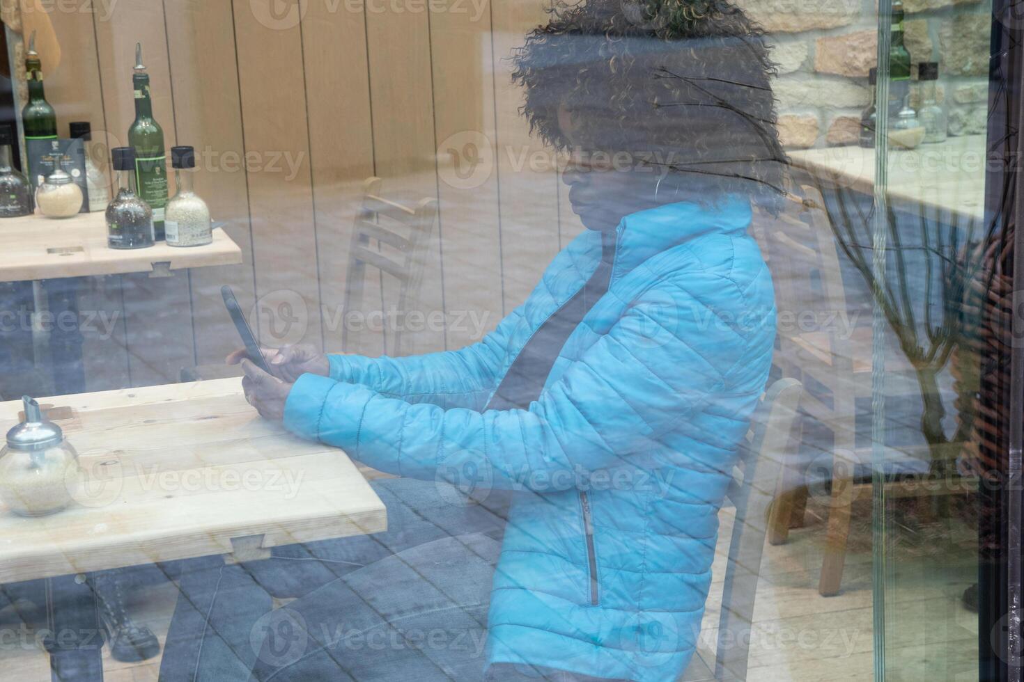 An African American woman in a blue jacket sits behind a window glass in a cafe makes an order using an application on a smartphone, selective soft focus from due to dirty glass. High quality photo