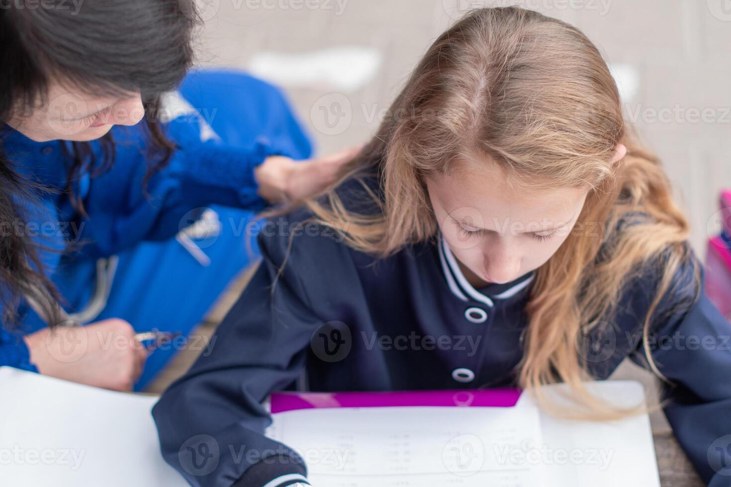 smart girl solving math problems on her own while her mother sits nearby at wooden table on children's playground and watches,education concept,going back to school, girl in beautiful blue uniform photo