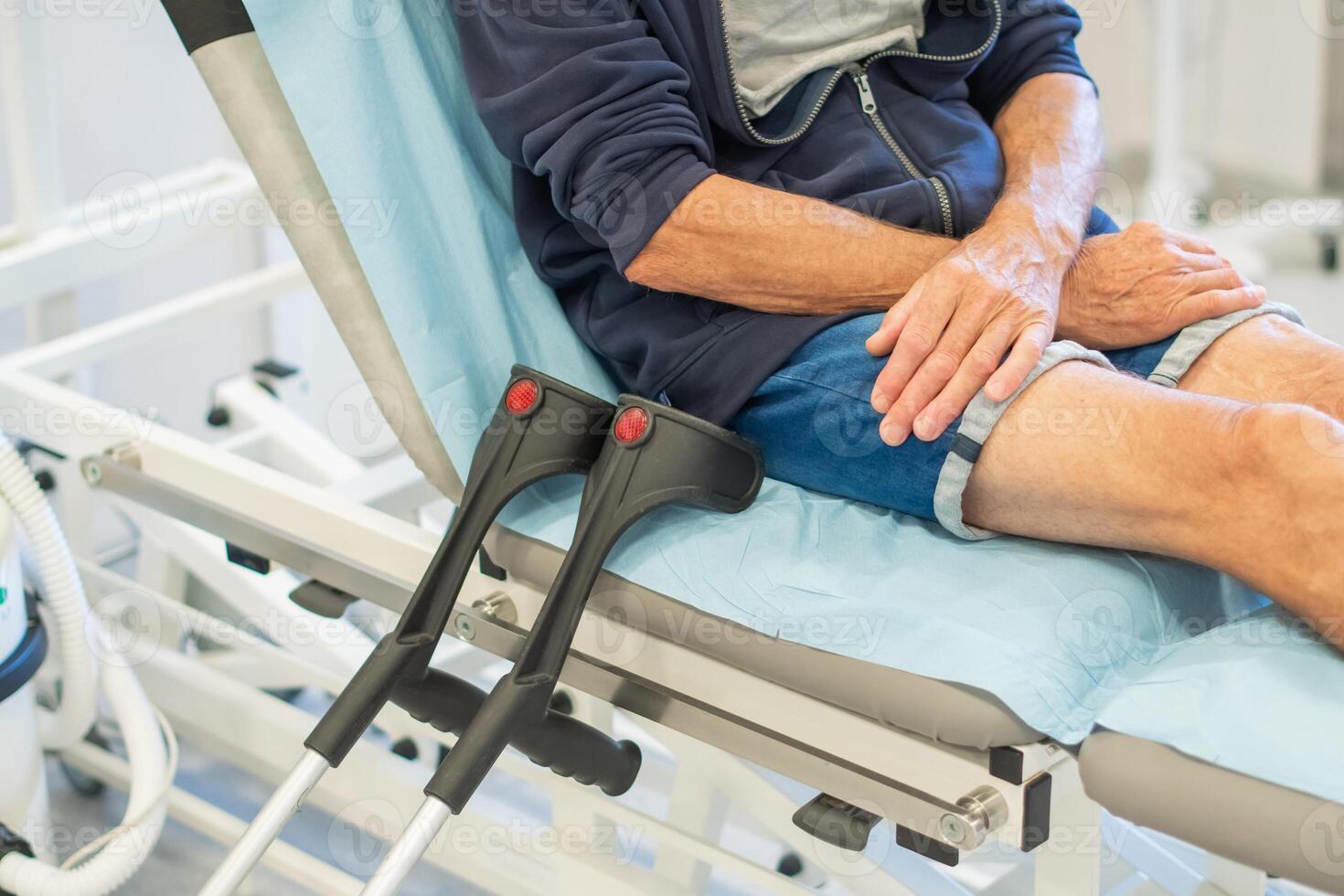 middle aged man sits deep in thought on a medical couch in a clinic office, waiting for the doctor to examine him and put a cast on his broken leg, his crutches lying next to the bed, photo
