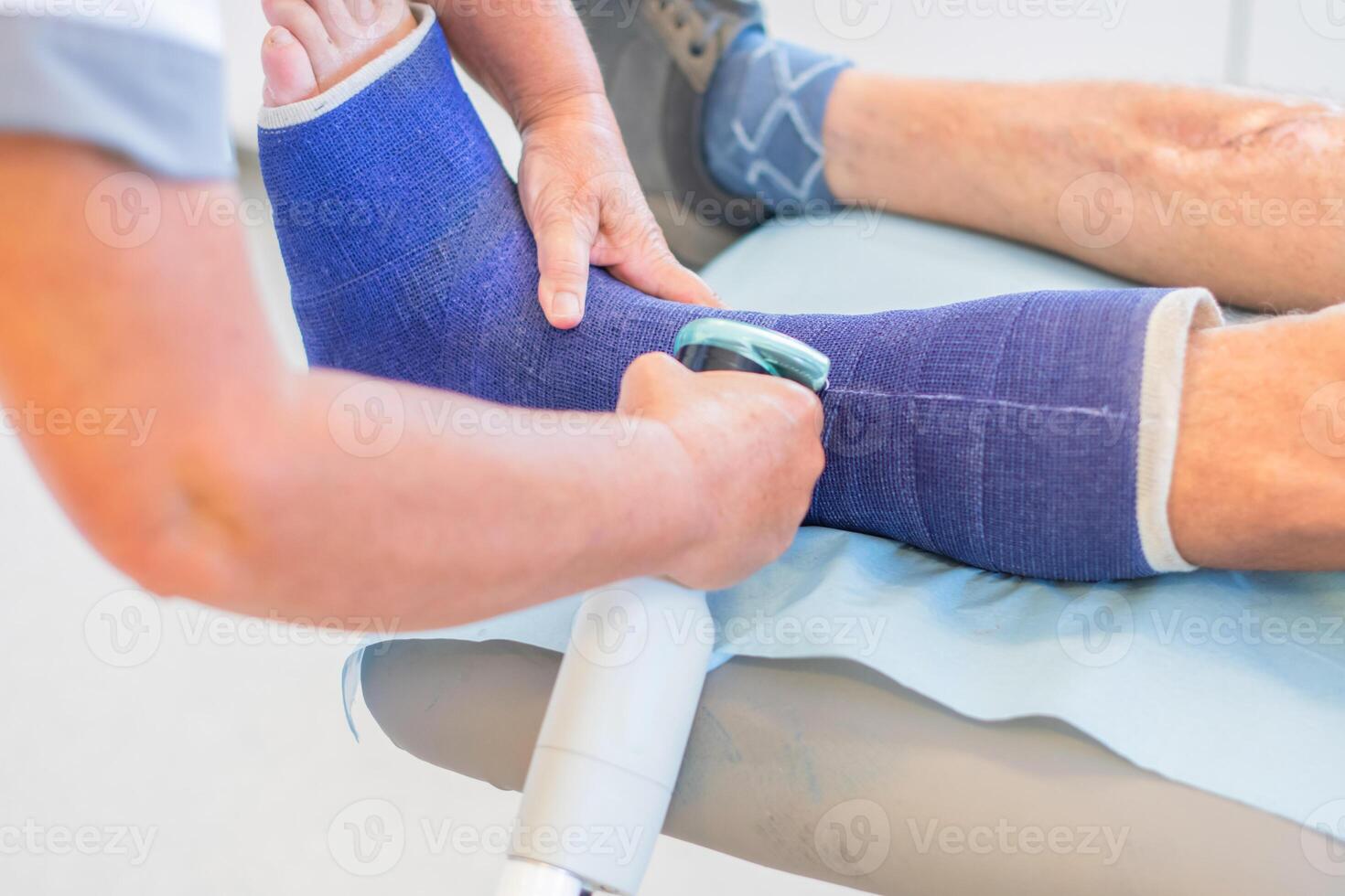 Nurse in clinic cutting strong fiberglass cast on patient's leg with plaster saw taking precautions, patient sitting quietly, professional help concept, photo