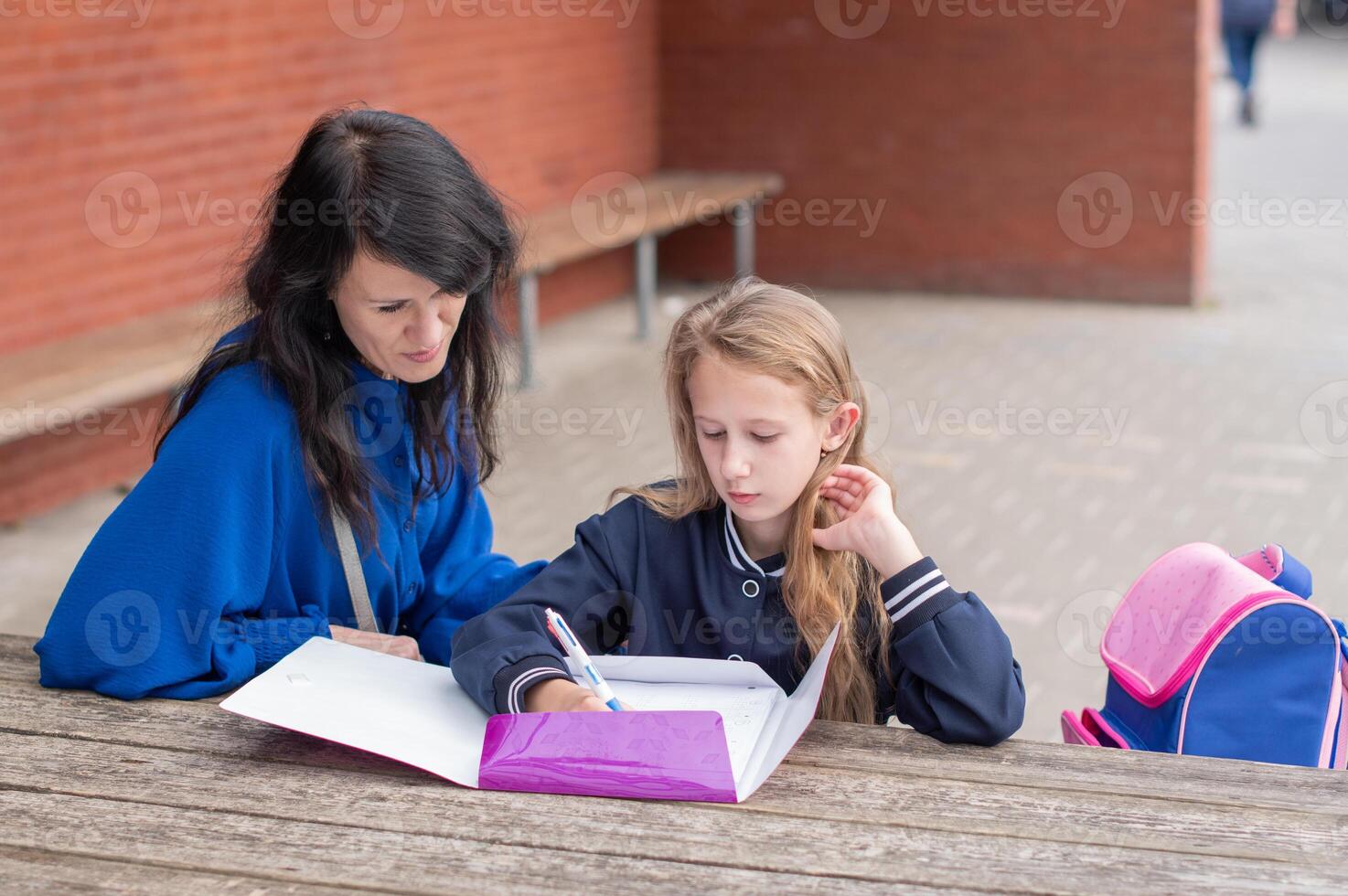 smart girl solving math problems on her own while her mother sits nearby at wooden table on children's playground and watches,education concept,going back to school, girl in beautiful blue uniform photo