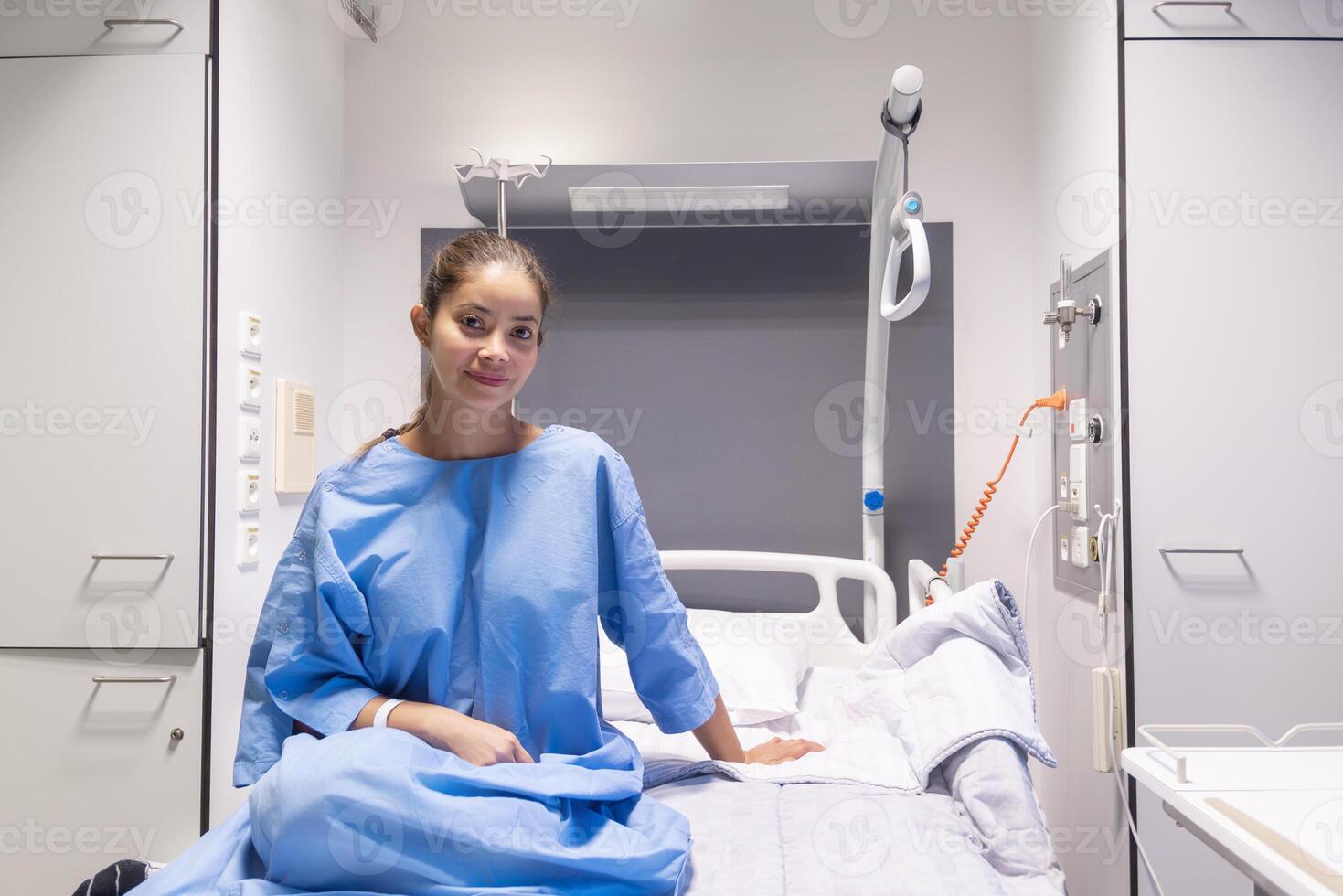 Young female patient in hospital ward,dressed in blue gown,sitting on bed and waiting to be examined by doctor,smiling softly and satisfied with service and services provided,individual hospital room photo