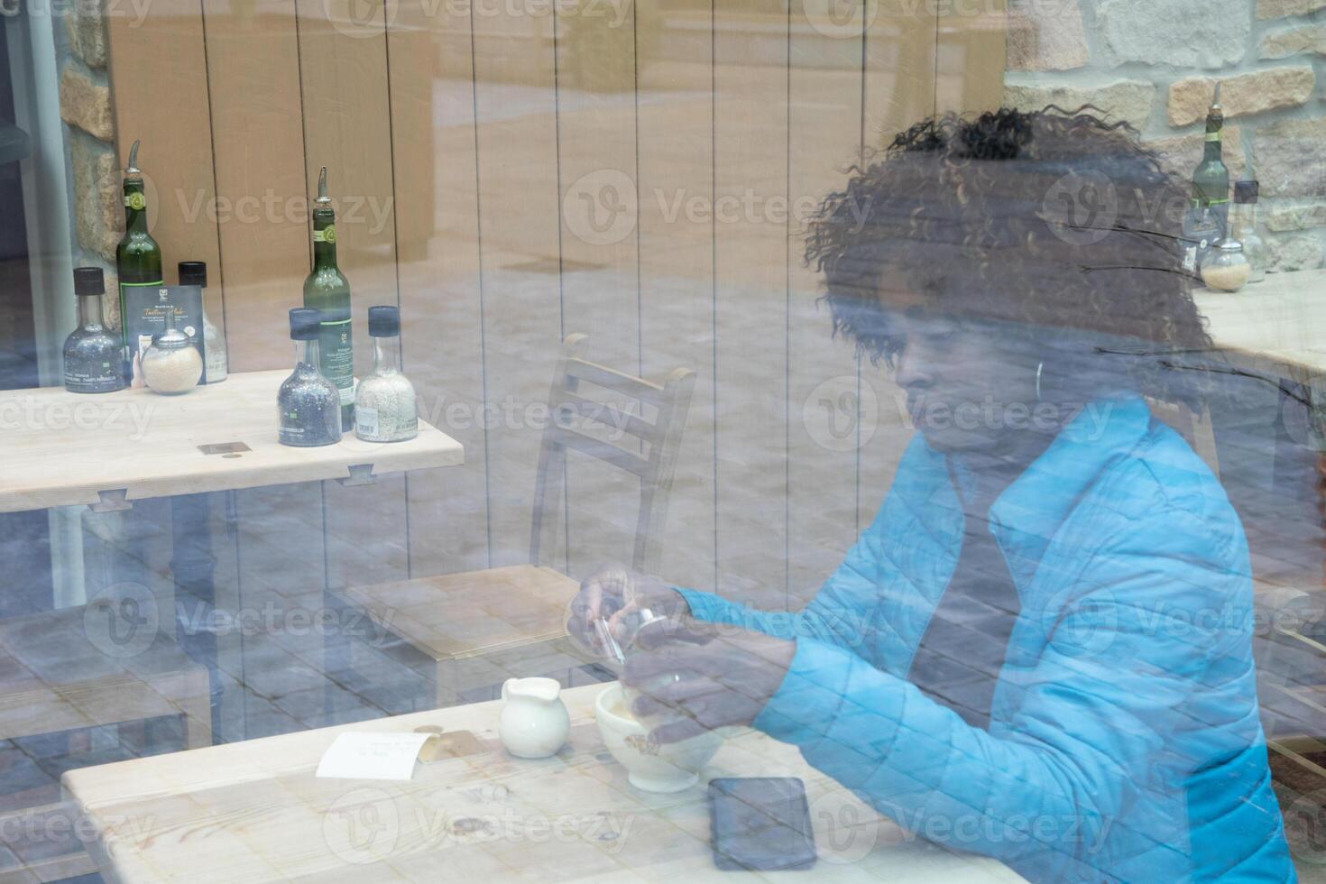 An African American woman in a blue jacket sits behind a window glass in a cafe makes an order using an application on a smartphone, selective soft focus from due to dirty glass. High quality photo