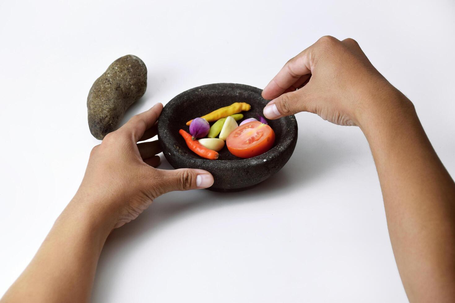 Two people's hands are making chili sauce using a mortar made of stone photo
