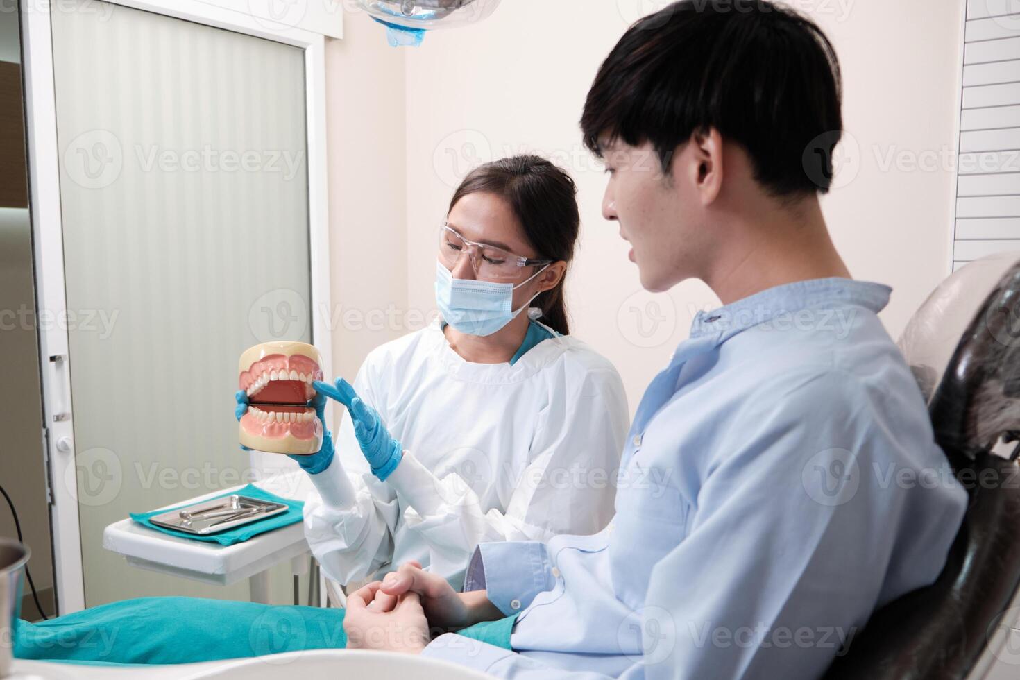 Asian female dentist explaining orthodontic care and whitening to young male patient with teeth model in dental clinic, well-being hygiene, and professional healthcare work in doctor office hospital. photo