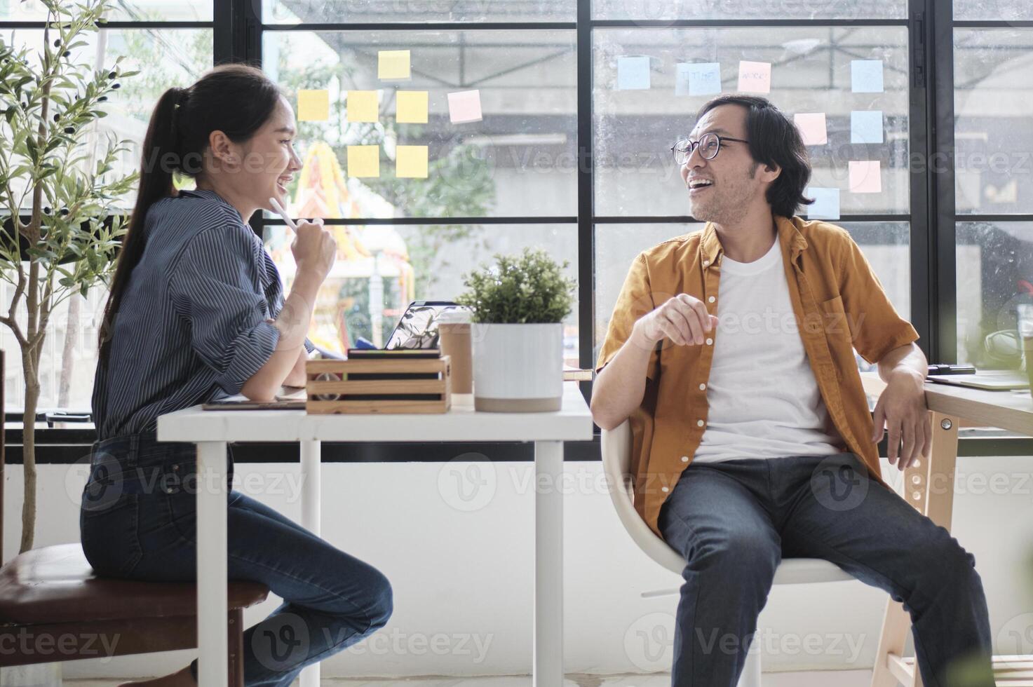 Two Asian workers sitting in chairs at working desks, collaborating and talking at daylight windows in casual small business office, online business job coworkers, positive relationship colleagues. photo