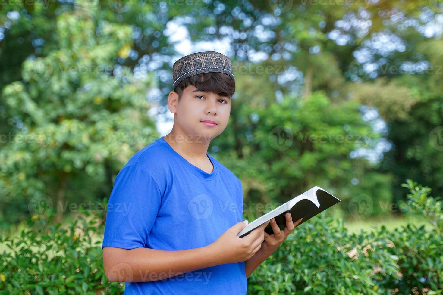 A young Muslim boy wearing a blue T-shirt and black Taqiyah cap poses for a photo reading a book in a park. Soft and selective focus.