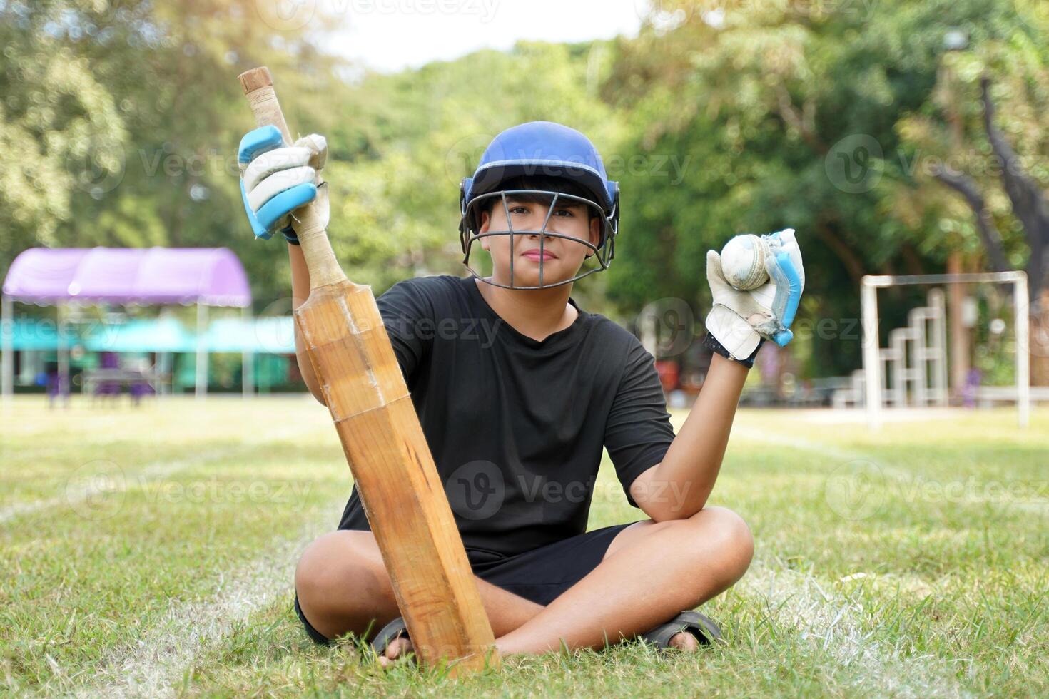 Cricket player wearing cricket Helmet holds a bat and ball in his hand, sitting on the grass while practicing. photo