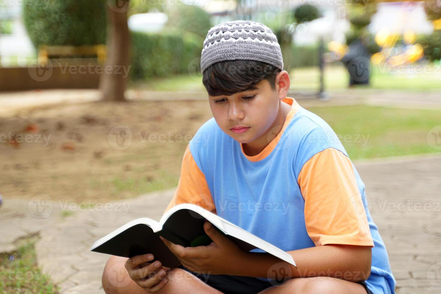 A young Muslim boy wears a blue T-shirt and a gray Taqiyah cap. Sitting and posing for photos while reading a book at the park.