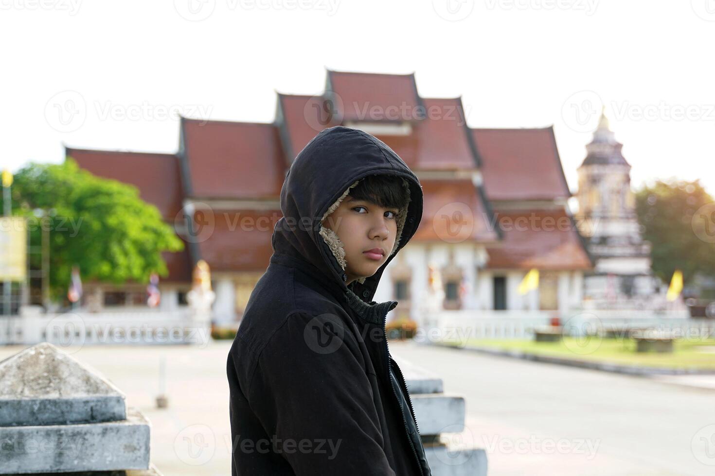 teenage boy with a hood covering his head stood alone in front of a Thai temple with no intention of going anywhere. concept of teenage problems, loneliness, homelessness. photo